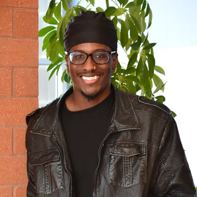 Link, Young man wearing a black leather jacket leans against a brick wall with plant in background, profile page